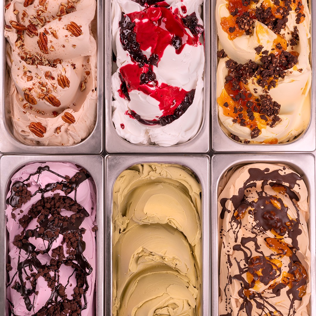 Six metal trays of gelato in different flavors, including pecan, berry, pistachio, and chocolate-caramel, viewed from...